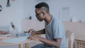 Investimento em Ações: Primeiros Passos: Homem sentado à mesa de trabalho em casa, concentrado enquanto usa seu laptop e escreve em papéis, possivelmente fazendo pesquisas ou estudando sobre investimentos.